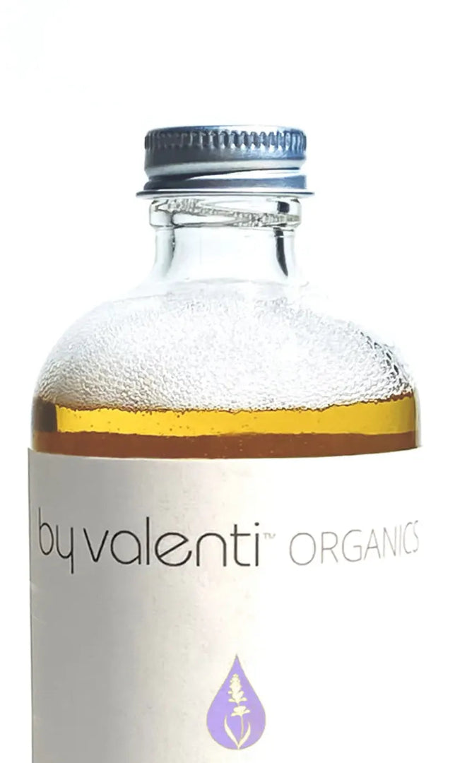 Close up of a BY VALENTI lavender and almond glass bottle of liquid soap with visible 'by valenti organics' branding on the label on a white background