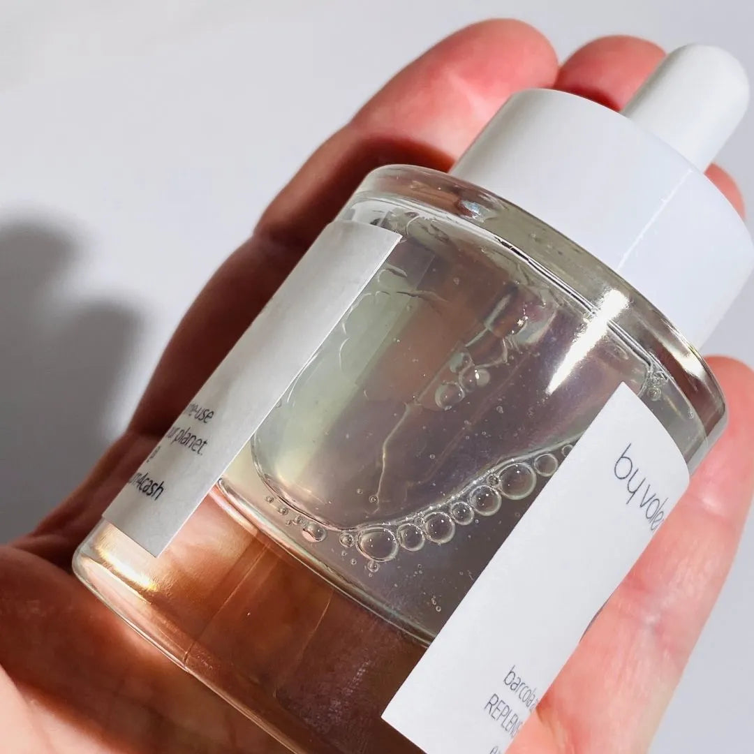 A hand holding a clear glass dropper bottle filled with BY VALENTI  Barcola Azzurro Mare Replenishing Elixir, featuring a white label and white dropper cap against a light background.