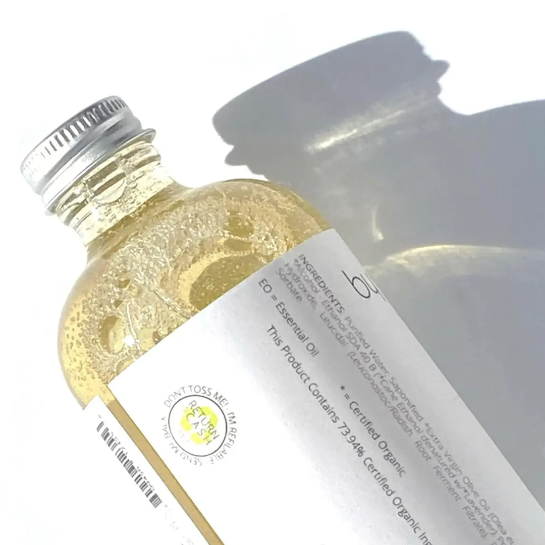 Close of back of the label of BY VALENTI Liquid Castile Soap in a glass clear bottle with an aluminium cap displaying their Return for Cash logo casting a long afternoon shadow on a white background