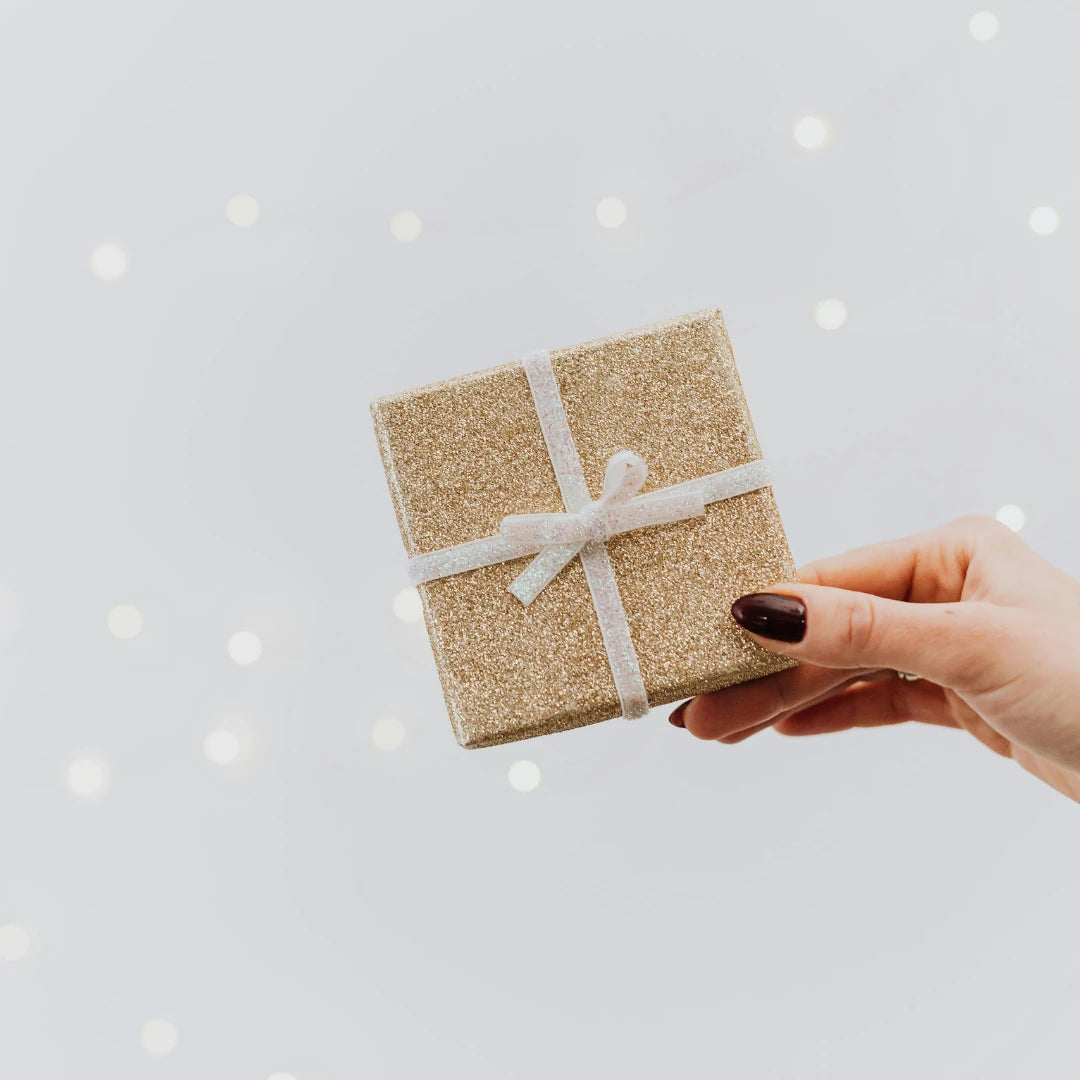 Hand holding a small glittery gift box with a white ribbon against a light gray background.