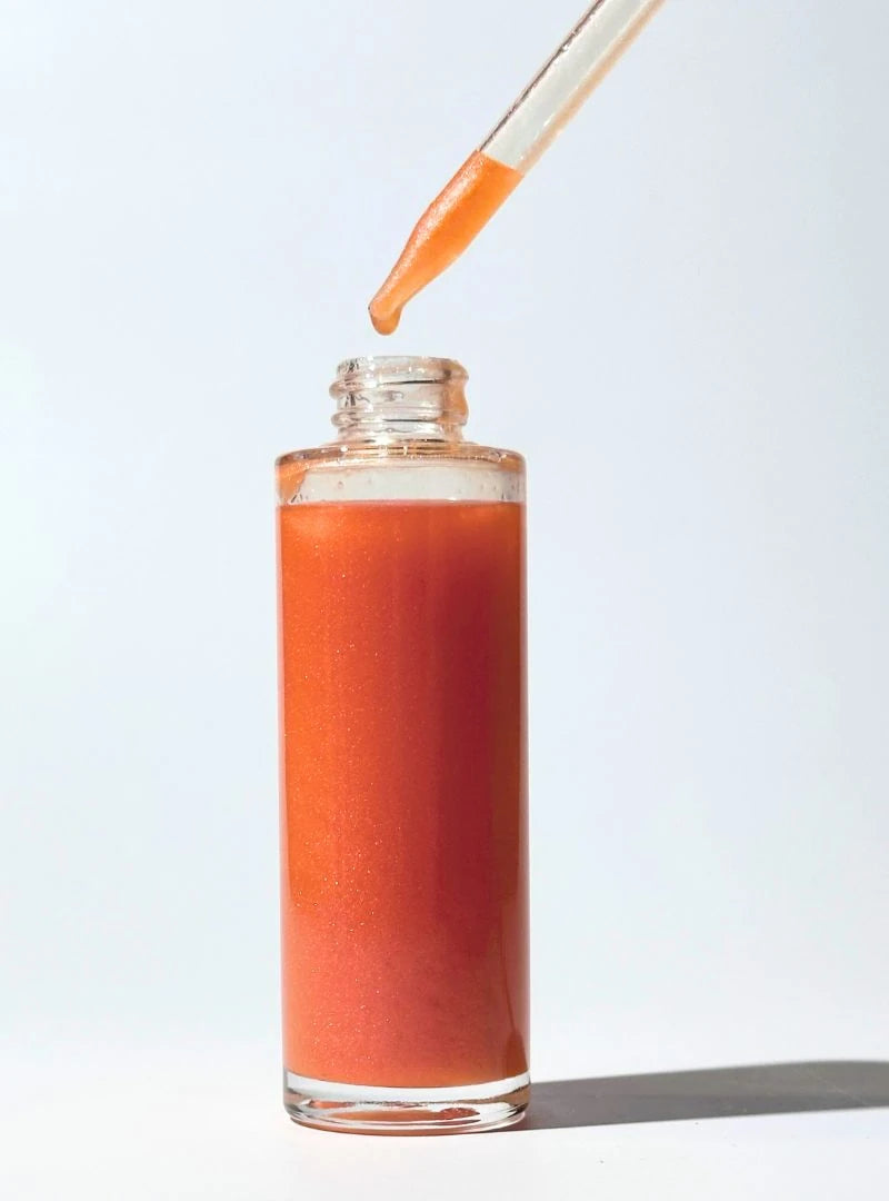 Glass bottle filled with dropper of By Valenti pink orange shimmering body oil being topped up with a dropper on a light gray background
