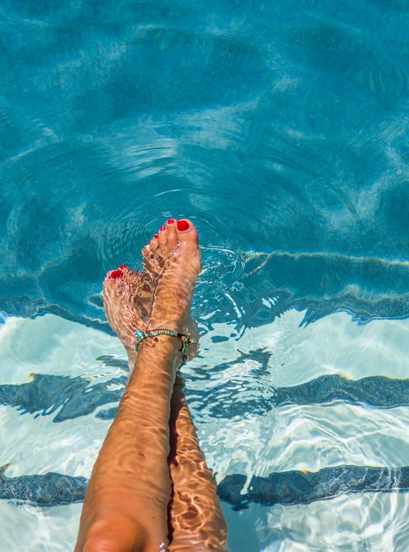 Woman feet with red nail polish and a bracelet in clear blue water pool