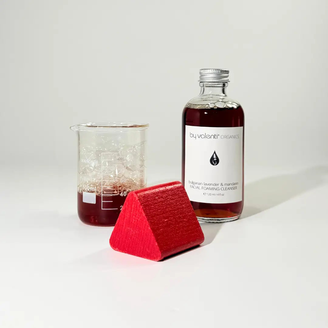 Glass bottle of naturally amber colored lavender facial soap with a beaker containing the same bubbly soap and a red red kids toy on a white background