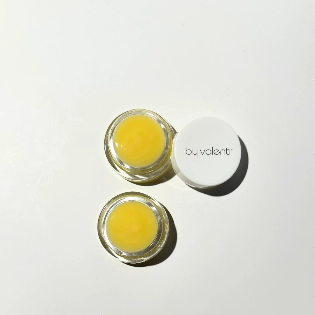 Two small jars of yellow eye balm with a white lid on a light gray background, branded 'by valenti'.